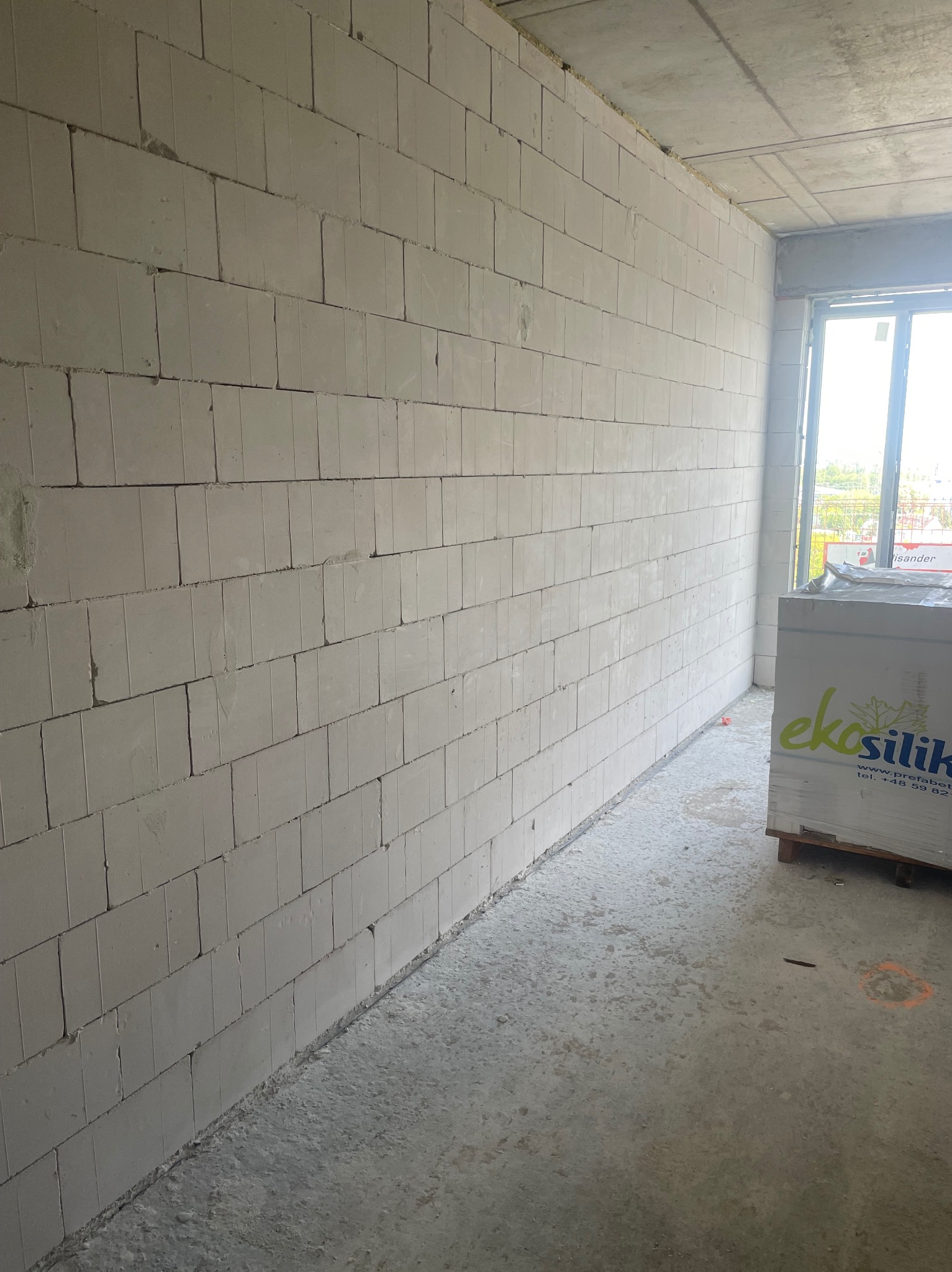 Wnętrze w trakcie budowy: ściana z białych bloczków silikatowych, paleta z materiałem budowlanym przy oknie. Widoczny surowy betonowy strop i podłoga.