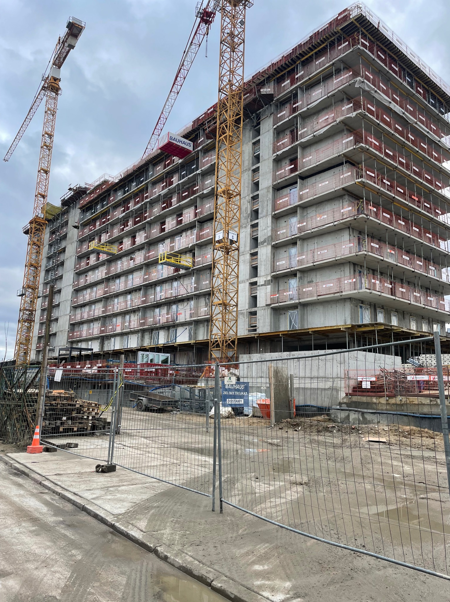Wielopiętrowy budynek w surowym stanie, z widocznymi żurawiami i elementami konstrukcyjnymi. Ujęcie z dołu, z widocznym ogrodzeniem placu budowy i błotnistym terenem.