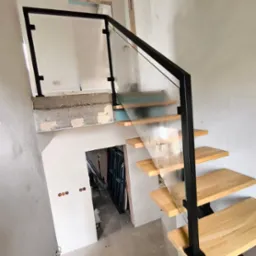 Nowoczesne schody z drewnianymi stopniami, czarną metalową balustradą i szklanym wypełnieniem, w surowym stanie wykończenia wnętrza.