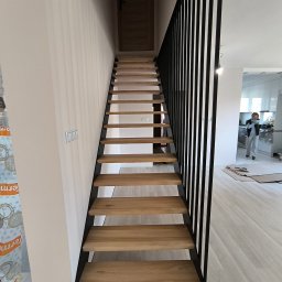 Stalove Tarnobrzeg Łukasz Załucki - Nowoczesne schody drewniane z czarną, pionową balustradą, widok z dołu, w tle osoba w kuchni.