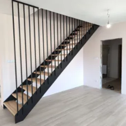 Schody drewniane z czarną metalową konstrukcją i pionowymi szczebelkami, widok z dołu na tle jasnego wnętrza w trakcie wykończenia.