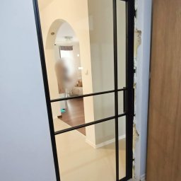 Stalove Tarnobrzeg Łukasz Załucki - Szklane drzwi z czarną metalową ramą, widoczne niedokończone wykończenie wokół ościeżnicy, wnętrze mieszkania w tle.