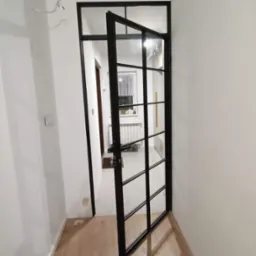 Wewnętrzne drzwi w stylu loftowym z czarną metalową ramą i szprosami, częściowo otwarte, widok na wnętrze mieszkania z grzejnikiem i oknem.