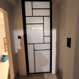 Stalove Tarnobrzeg Łukasz Załucki - Nowoczesna, wewnętrzna, przeszklona konstrukcja z czarnymi profilami, dzieląca przestrzeń w pomieszczeniu o beżowych ścianach i ceglanej podłodze.