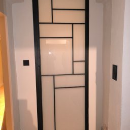 Stalove Tarnobrzeg Łukasz Załucki - Czarna, metalowa konstrukcja drzwi z mlecznymi szybami w geometryczne wzory, osadzona w białej ścianie nad ceglaną podłogą.