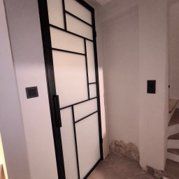Stalove Tarnobrzeg Łukasz Załucki - Nowoczesne drzwi wewnętrzne z czarną ramą i białymi panelami w geometryczne wzory, zamontowane w surowym wnętrzu z widoczną instalacją elektryczną i fragmentem schodów w tle.