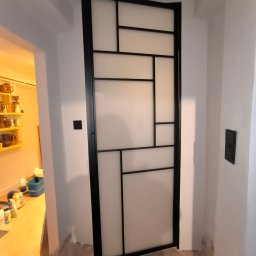 Stalove Tarnobrzeg Łukasz Załucki - Metalowa konstrukcja z mlecznym wypełnieniem, tworząca geometryczny wzór w czarnej ramie, prawdopodobnie drzwi lub ścianka działowa, w jasnym wnętrzu z terakotową podłogą.