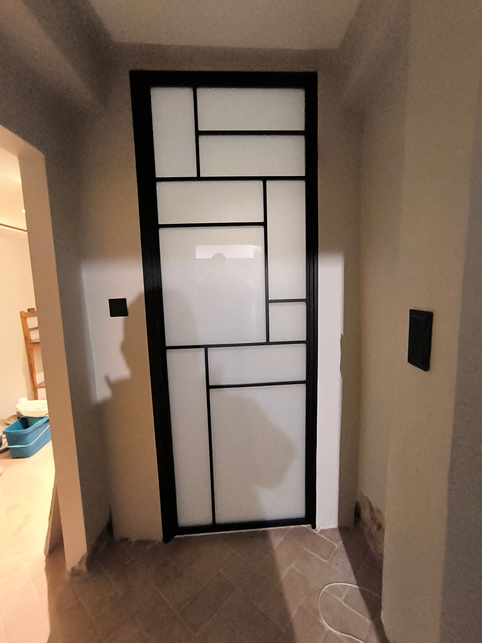 Nowoczesna, wewnętrzna, przeszklona konstrukcja z czarnymi profilami, dzieląca przestrzeń w pomieszczeniu o beżowych ścianach i ceglanej podłodze.