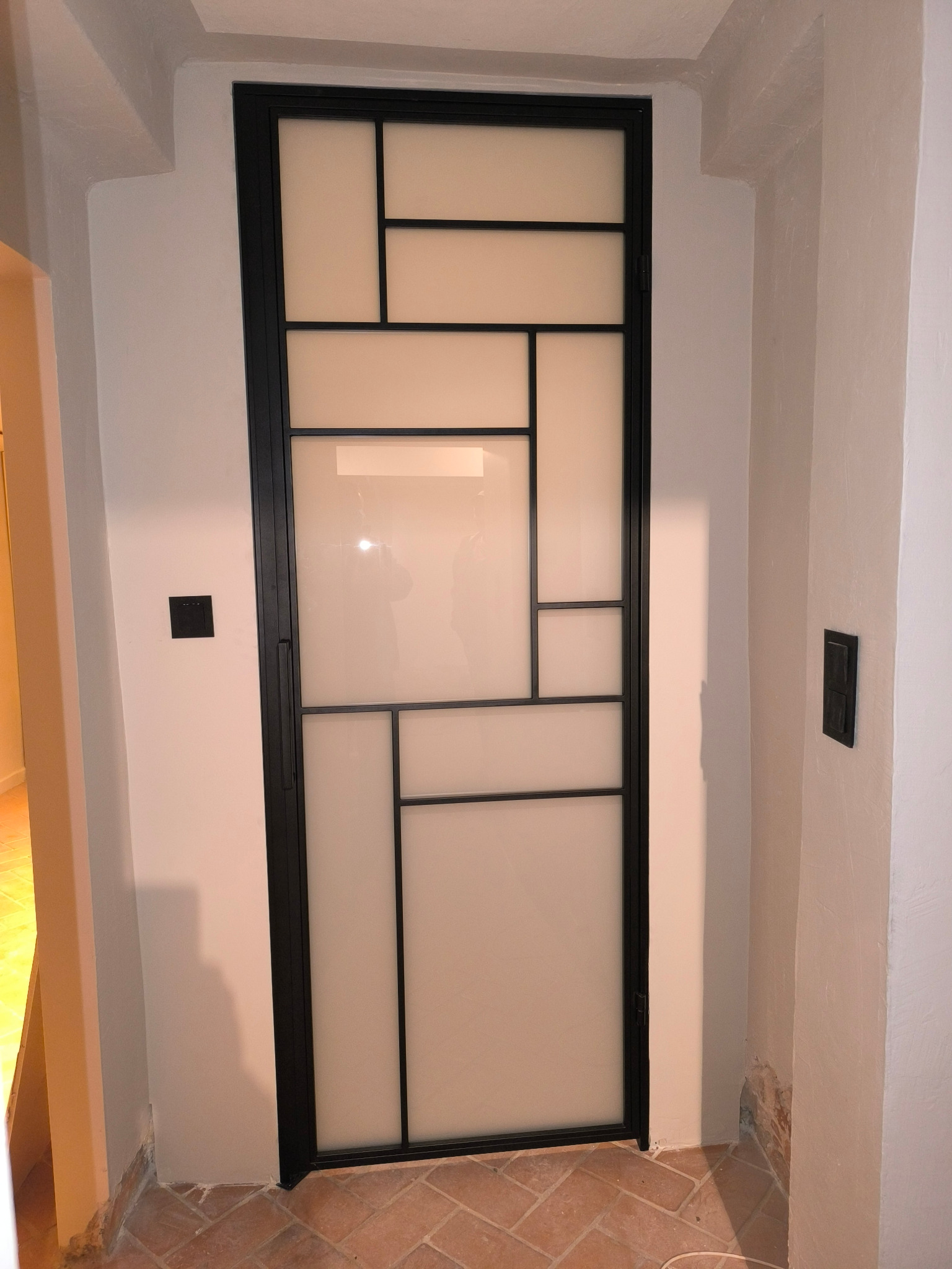 Czarna, metalowa konstrukcja drzwi z mlecznymi szybami w geometryczne wzory, osadzona w białej ścianie nad ceglaną podłogą.