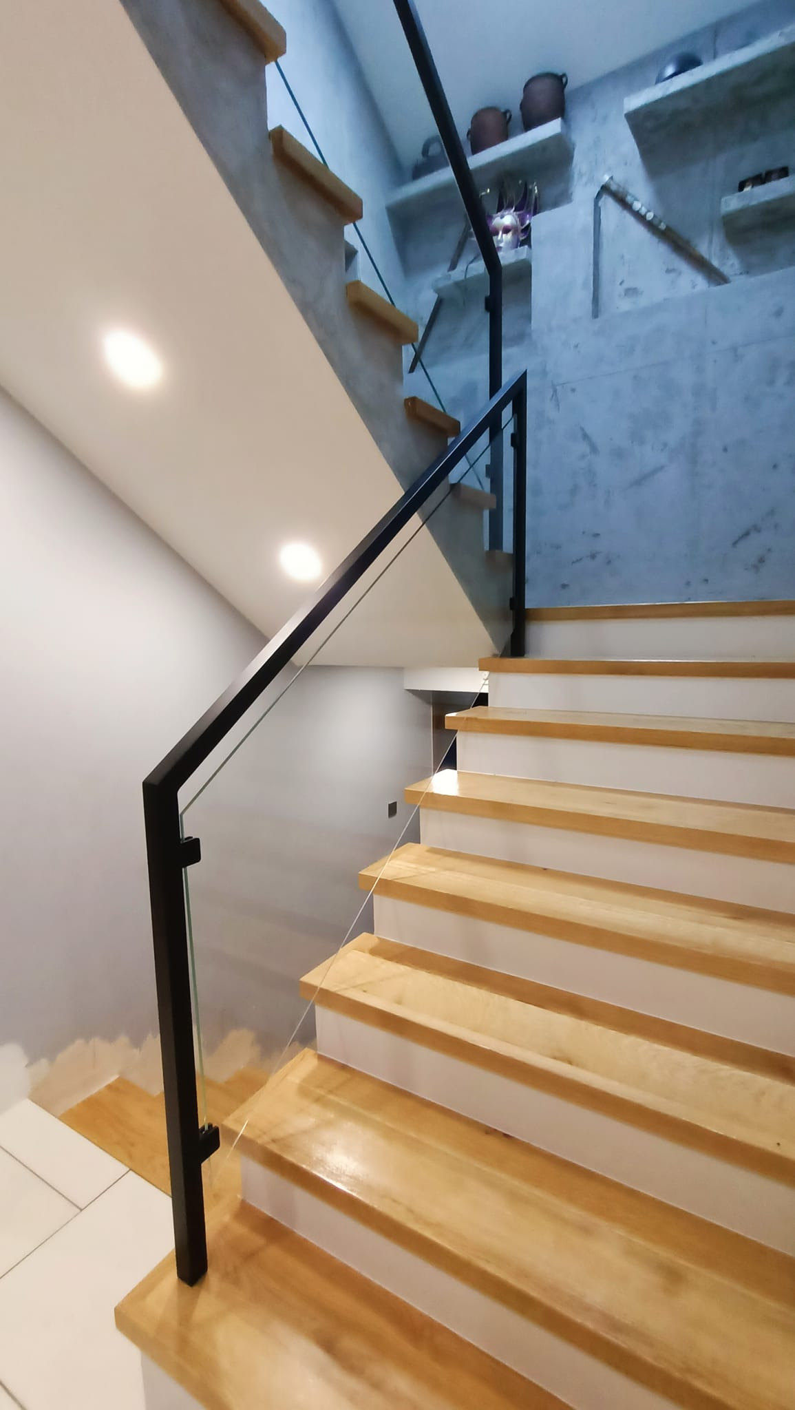 Schody drewniane z czarną balustradą szklaną, widok z dołu, minimalistyczny design, dekoracyjne półki z ceramiką w tle.