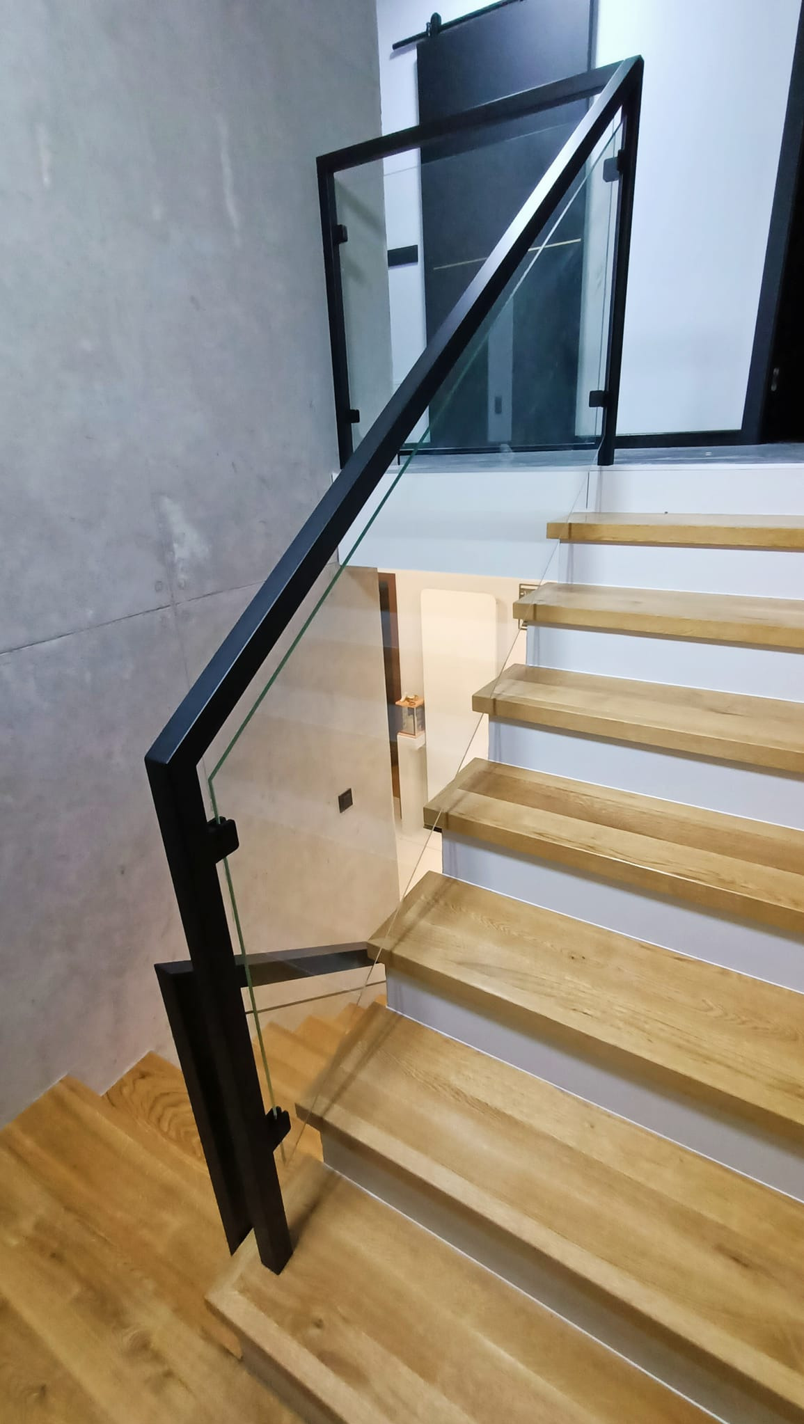 Szklana balustrada z czarnymi elementami mocującymi na drewnianych schodach w nowoczesnym wnętrzu.