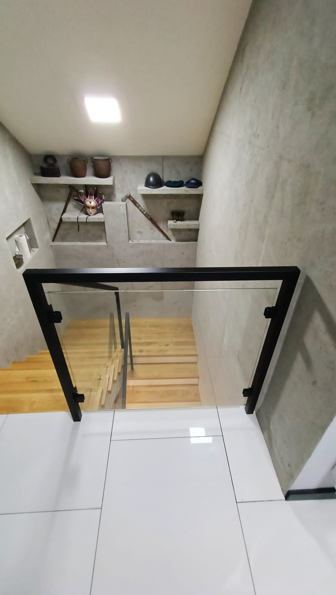 Szklana balustrada z czarnymi profilami zabezpieczająca schody z jasnego drewna, widok z góry na białe płytki podłogowe, ściany z betonu architektonicznego, na półkach dekoracje.