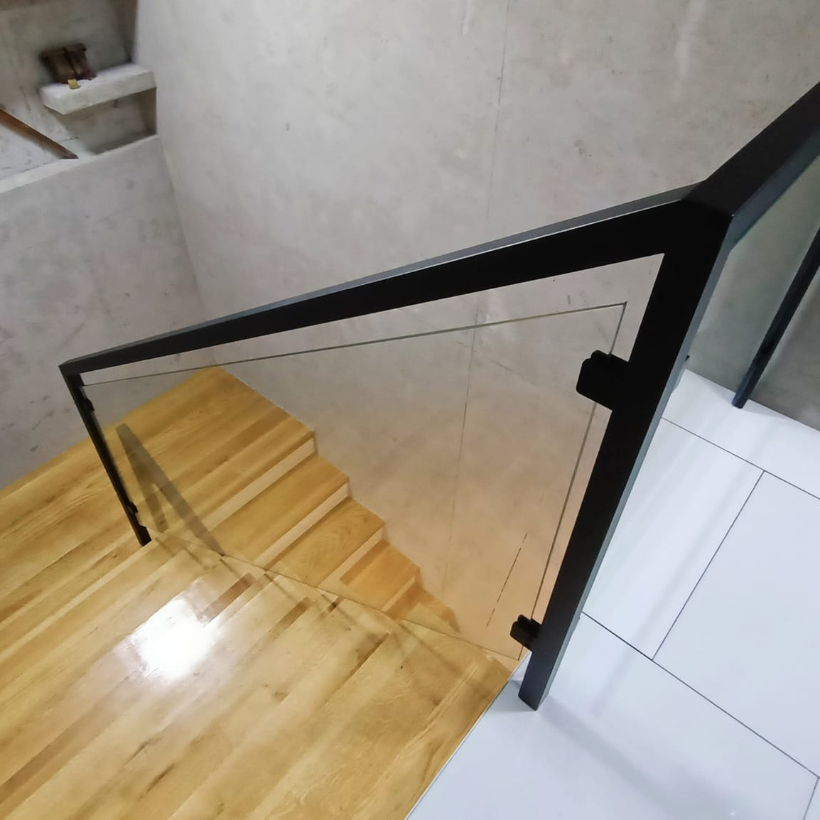 Szklana balustrada z czarną ramą zamontowana przy drewnianych schodach, widok z góry, białe płytki podłogowe w tle.