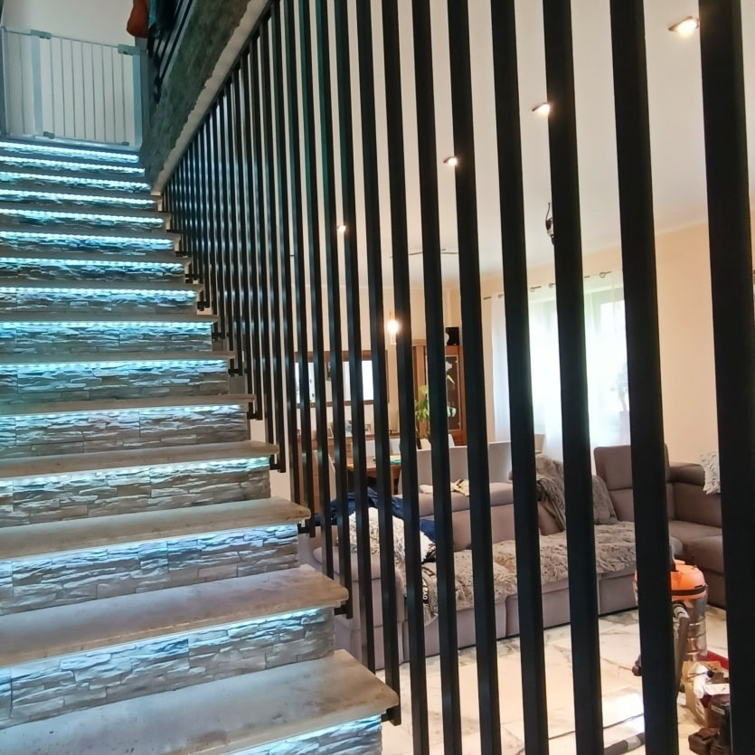 Nowoczesna klatka schodowa z podświetlanymi stopniami i czarną, pionową balustradą z metalowych prętów, widok na salon z szarą sofą.