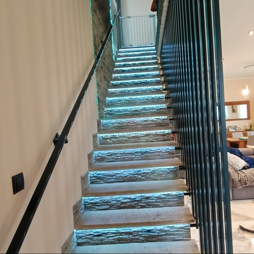 Schody wewnętrzne z podświetleniem LED, wykończone kamieniem dekoracyjnym, z czarną poręczą i ażurową ścianką działową.