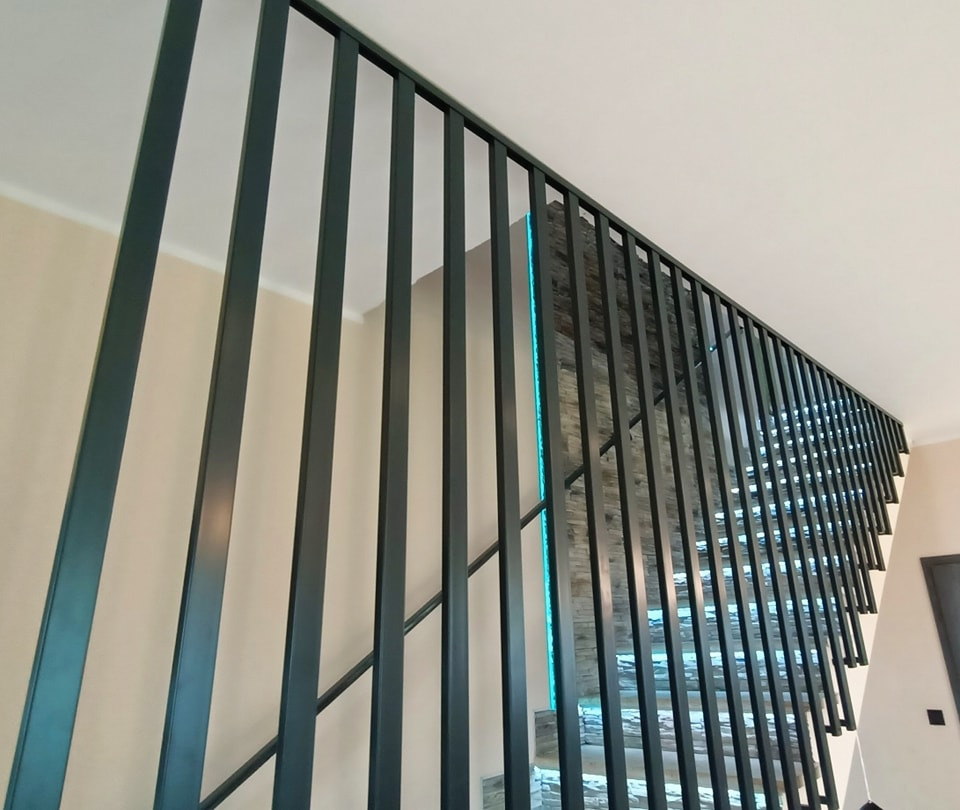 Nowoczesna balustrada schodowa z pionowych, metalowych profili w kolorze grafitowym, z podświetleniem LED na ścianie obłożonej kamieniem dekoracyjnym.