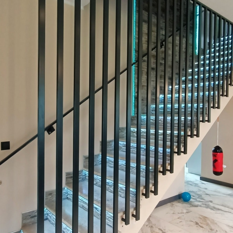Nowoczesne schody z czarną, pionową balustradą i podświetlanymi stopniami, widoczne z boku, z elementami dekoracyjnymi w tle.