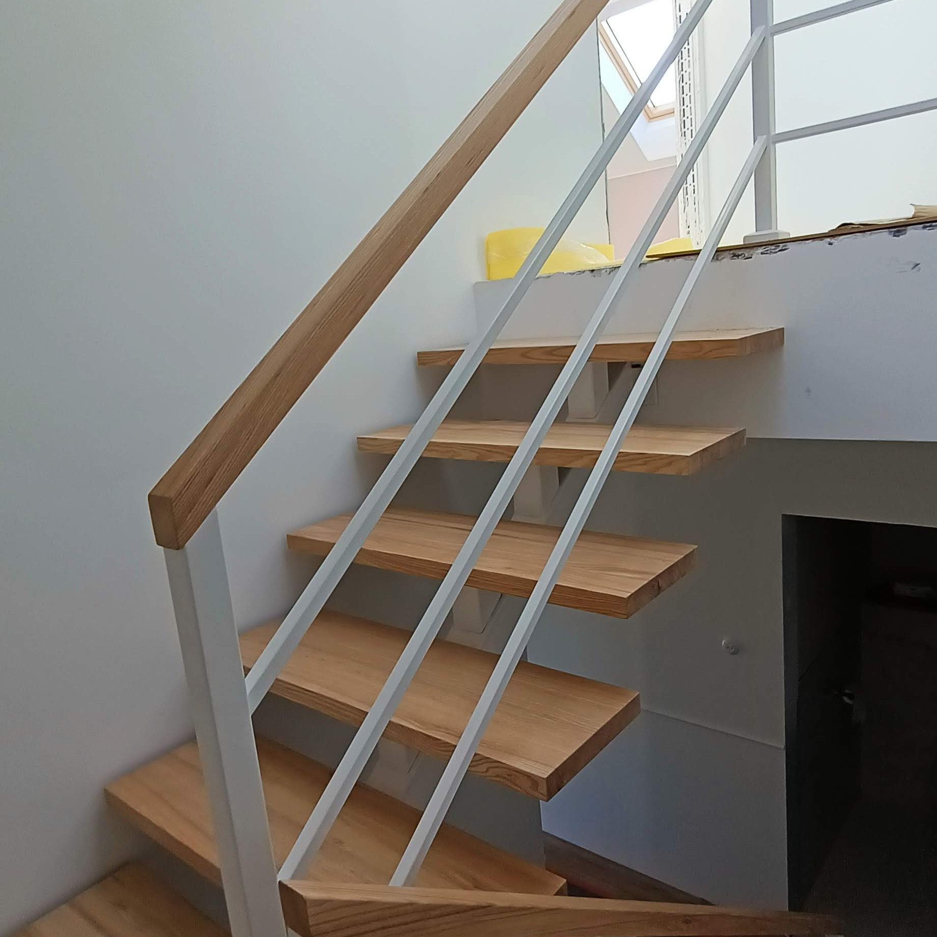 Schody drewniane z białą balustradą i drewnianą poręczą, widoczne z dołu, z oknem dachowym w tle.