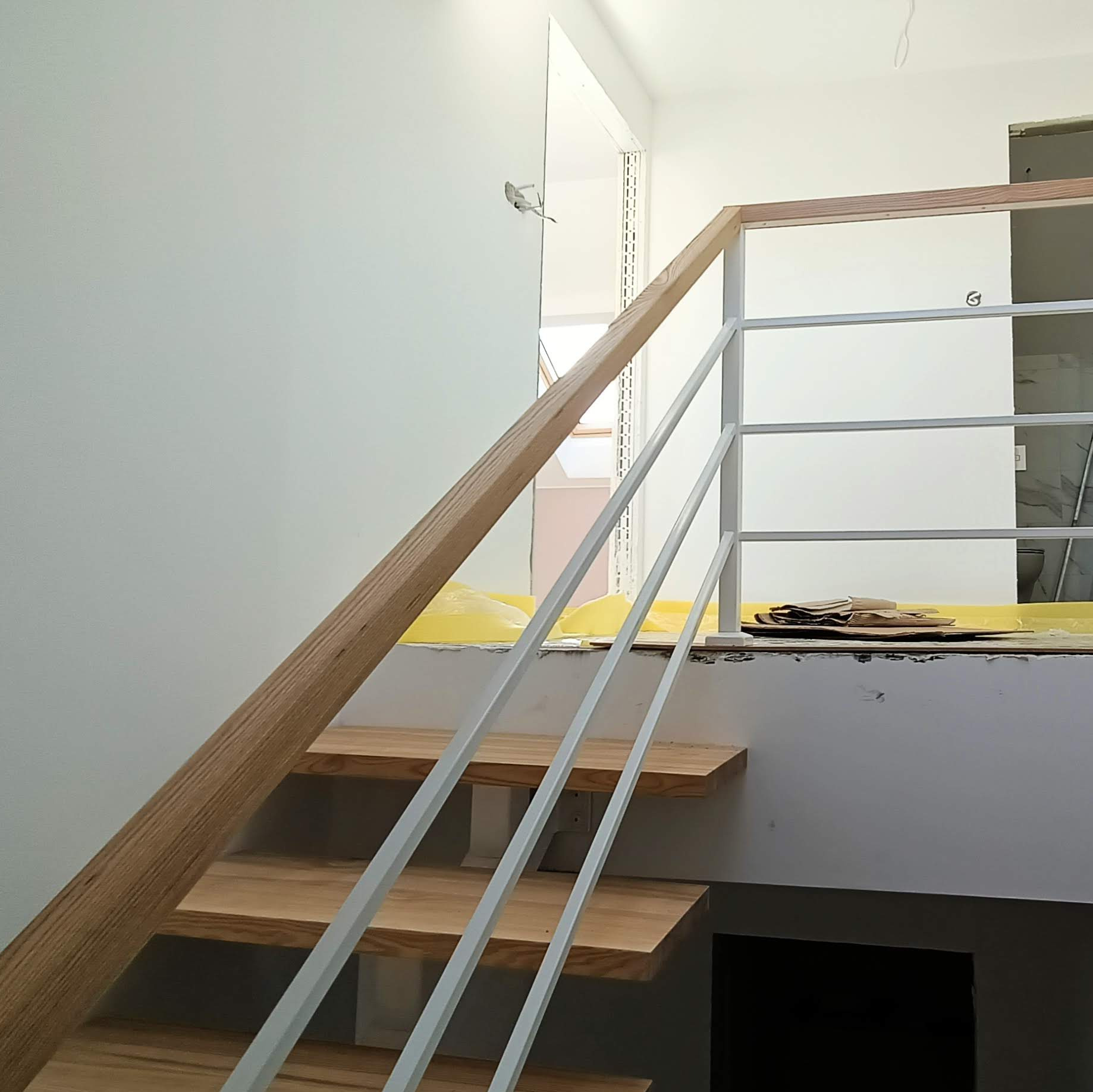 Wnętrze w trakcie wykańczania z drewnianymi schodami i białą balustradą stalową, widoczne zabezpieczenia podłogi i fragment łazienki w tle.