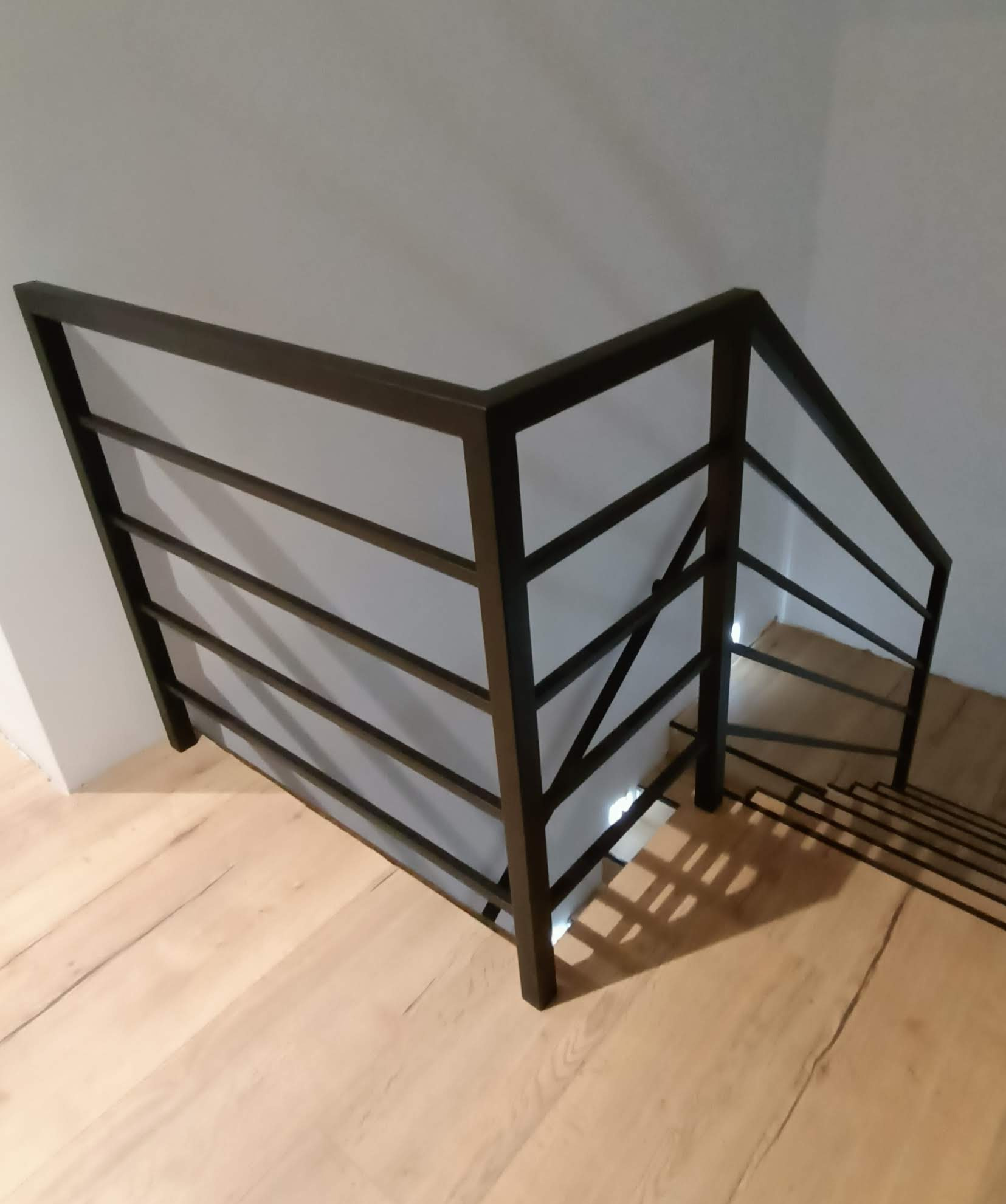 Minimalistyczna, czarna balustrada schodowa z poziomych profili na drewnianych schodach i białej ścianie, widok z góry.