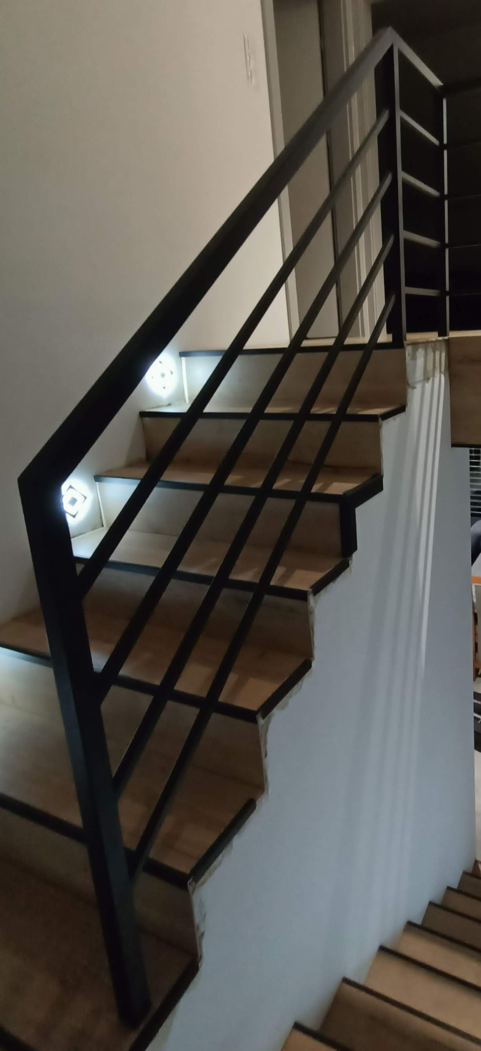 Schody z drewnianymi stopniami i czarną, metalową balustradą, podświetlone punktowymi lampkami LED wmontowanymi w stopnie, widoczne niedokończone wykończenie ściany.