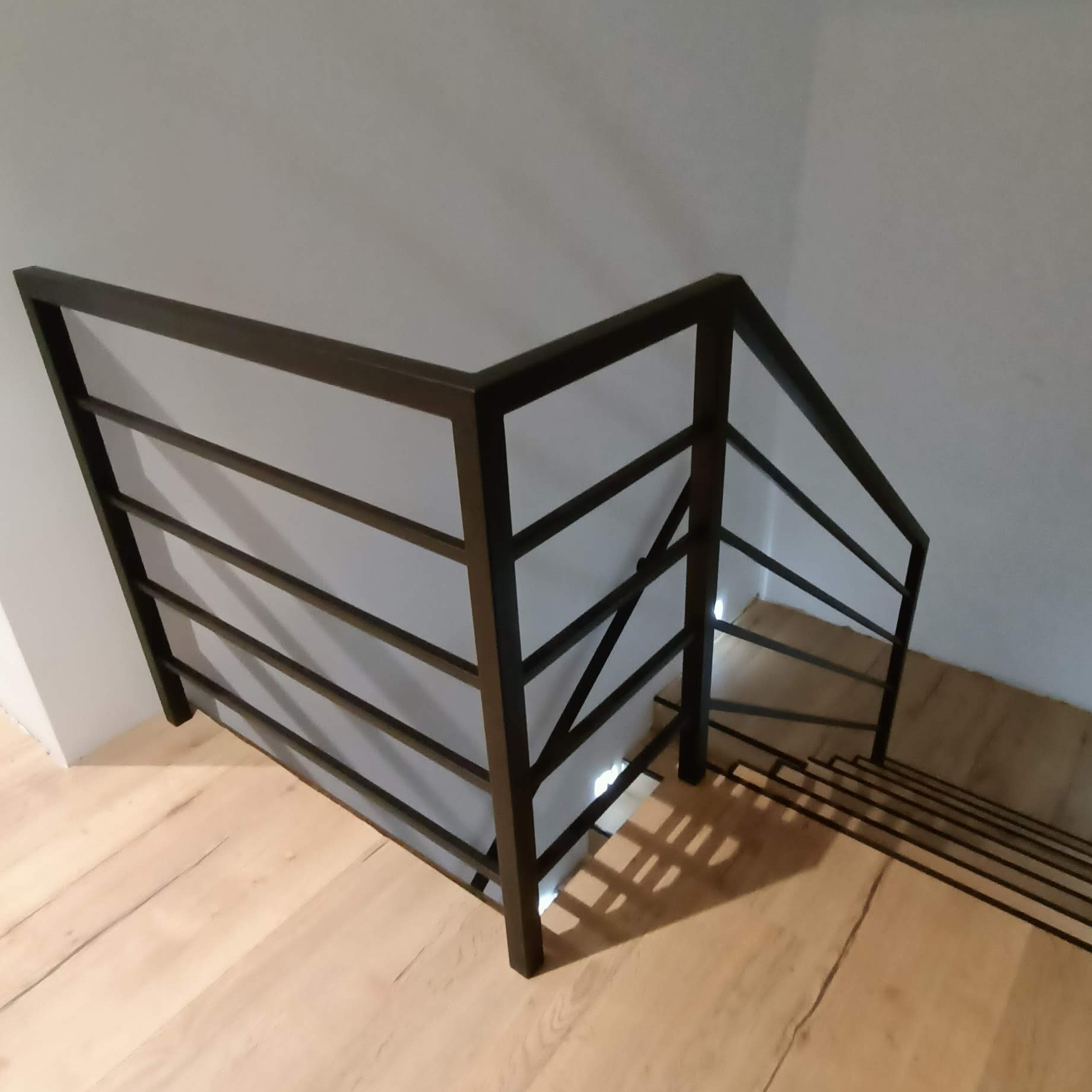 Minimalistyczna, czarna, metalowa barierka schodowa z poziomymi prętami, zamontowana w jasnym wnętrzu na drewnianych schodach, widok z góry.