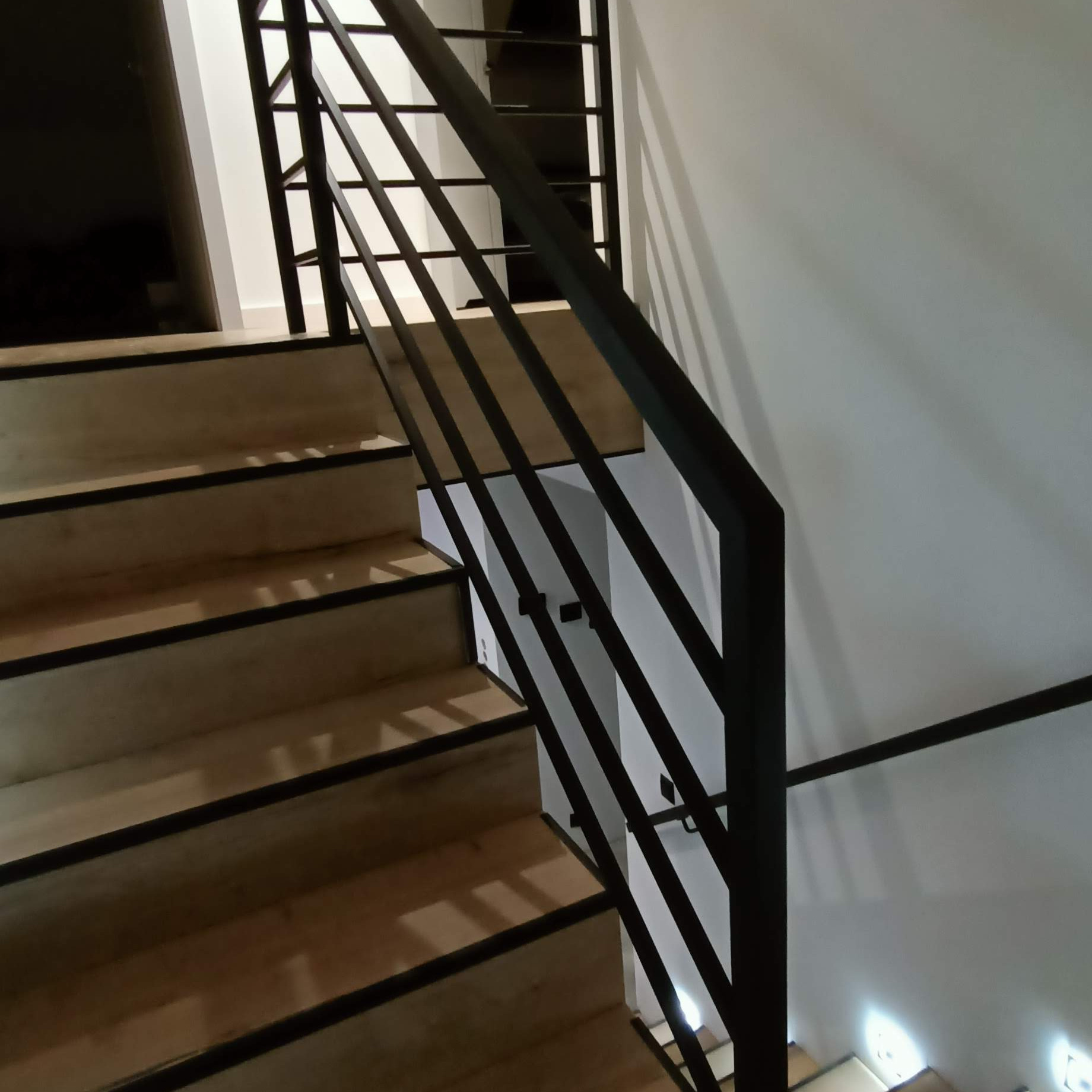 Minimalistyczna, czarna poręcz schodowa z prostych, pionowych prętów, zamontowana przy jasnych, drewnianych schodach w nowoczesnym wnętrzu.