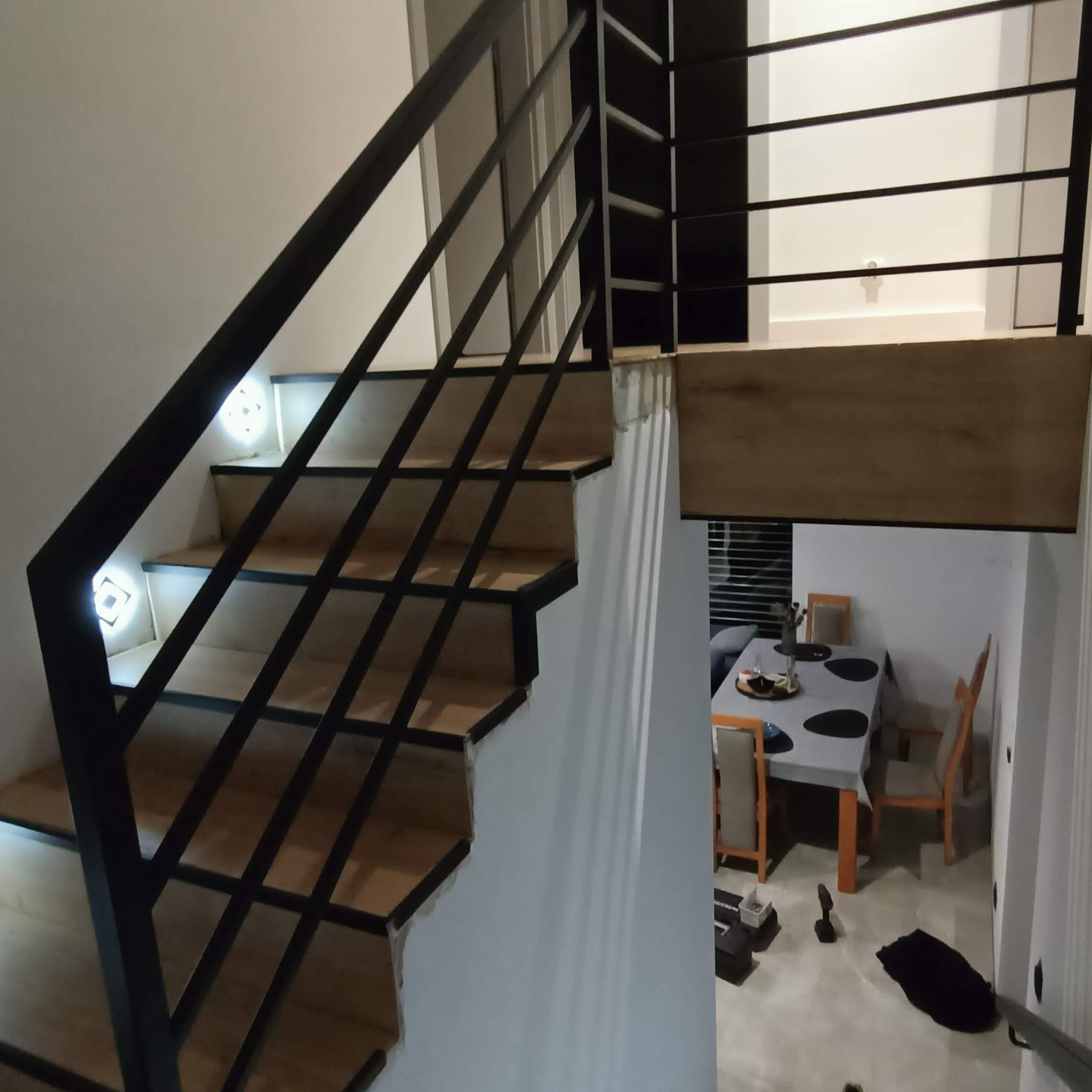 Schody wewnętrzne z drewnianymi stopniami i czarną, metalową balustradą, oświetlone punktowymi lampami w ścianie, z widokiem na jadalnię w tle.