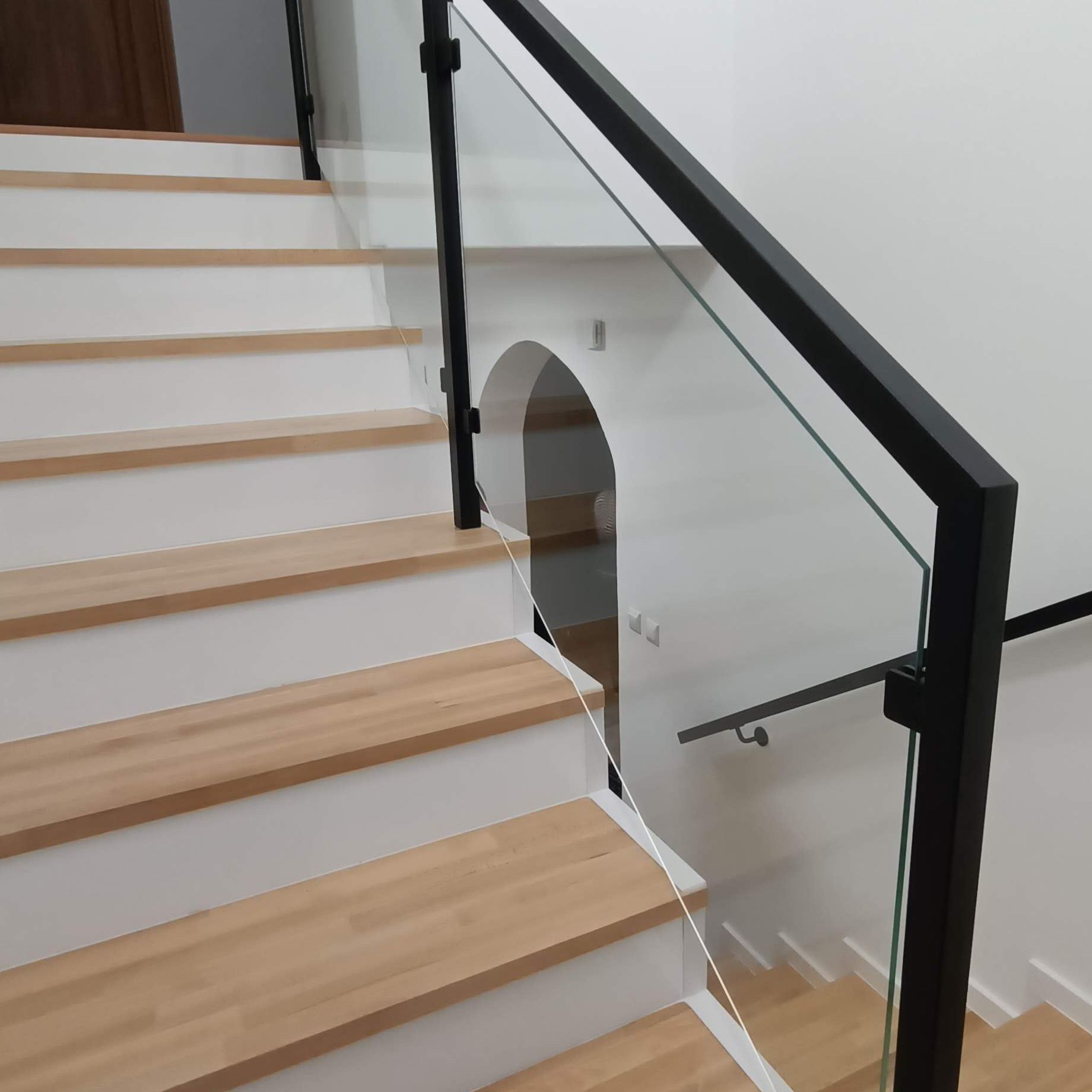 Nowoczesne schody z drewnianymi stopniami i szklaną balustradą z czarnymi, metalowymi słupkami i poręczą. Widoczny fragment łukowatego przejścia w ścianie.