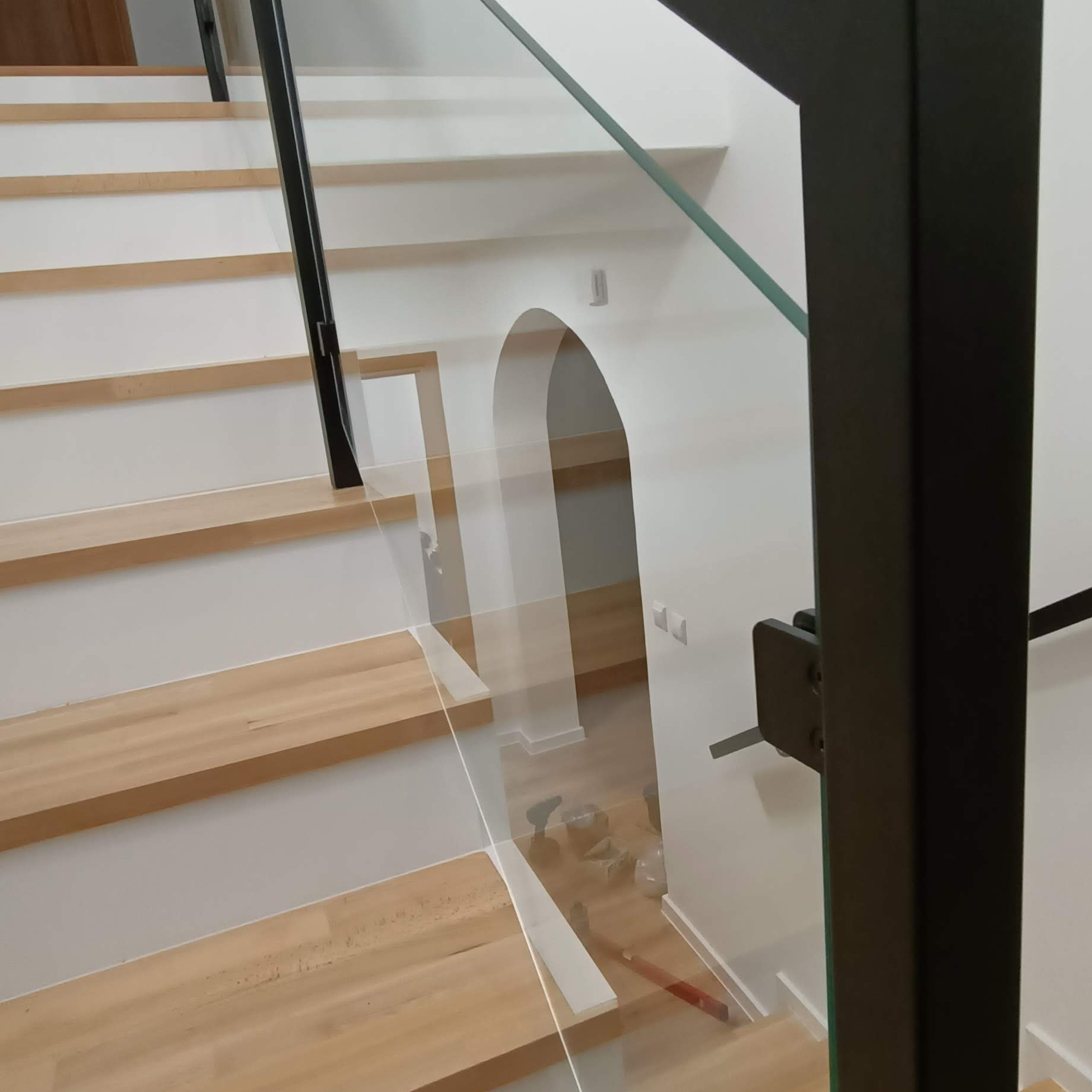 Szklana balustrada z czarnymi elementami mocującymi, zamontowana przy drewnianych schodach w jasnym wnętrzu z łukowym przejściem.