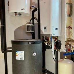 SANITECH Szymon Perkowski - Instalacja kotła gazowego Midea, zbiornika buforowego Aquatec Systems i zasobnika Galmet w kotłowni. Widoczne rury instalacyjne, zawory, manometry i skrzynka elektryczna na ścianie.