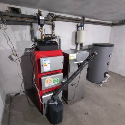 SANITECH Szymon Perkowski - Czerwony piec CO marki Kotlostal z podajnikiem, zbiornikiem na wodę i elementami instalacji grzewczej w szarej kotłowni.