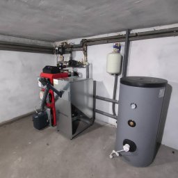SANITECH Szymon Perkowski - Zainstalowany piec CO marki KOTŁOSTAL z szarym zasobnikiem ciepłej wody w piwnicy z widocznymi rurami grzewczymi pod sufitem.