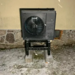 Zewnętrzna jednostka pompy ciepła Panasonic Aquarea zamontowana na metalowej konstrukcji, stojącej na betonowych bloczkach, w zimowej scenerii z padającym śniegiem, na tle ściany budynku...