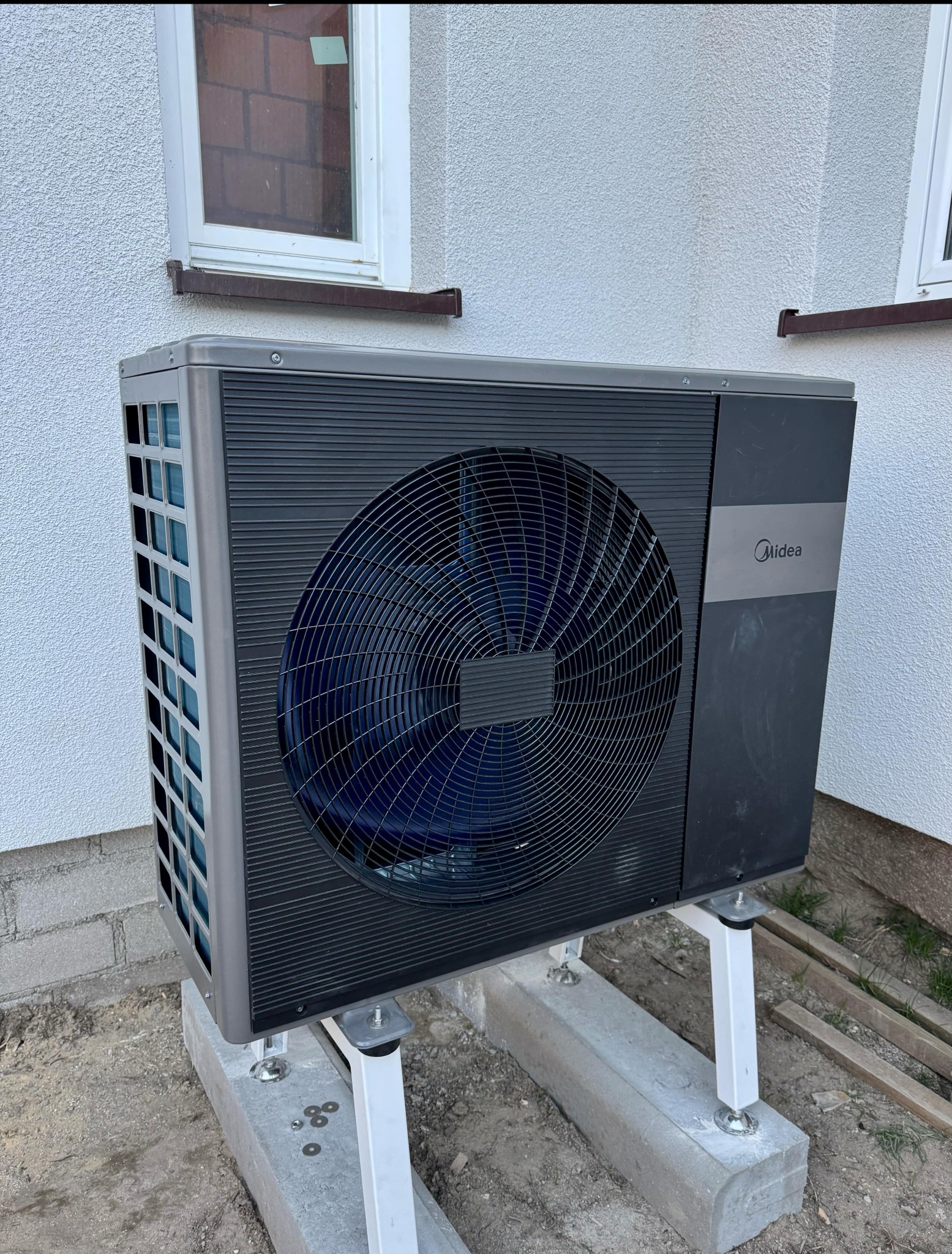 Zewnętrzna jednostka pompy ciepła Midea zamontowana na betonowych blokach z białymi wspornikami przy ścianie budynku z oknem.