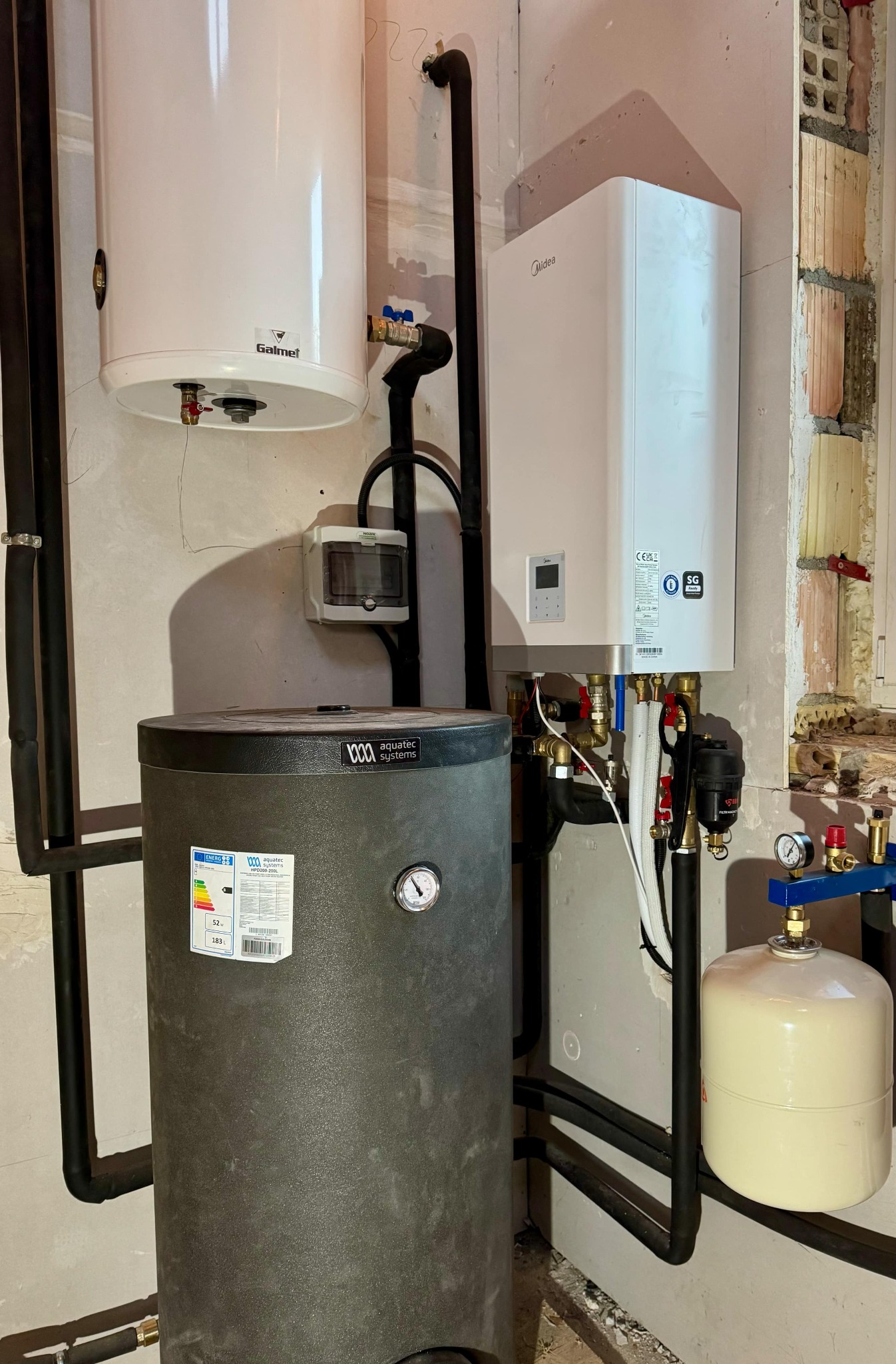 Instalacja kotła gazowego Midea, zbiornika buforowego Aquatec Systems i zasobnika Galmet w kotłowni. Widoczne rury instalacyjne, zawory, manometry i skrzynka elektryczna na ścianie.