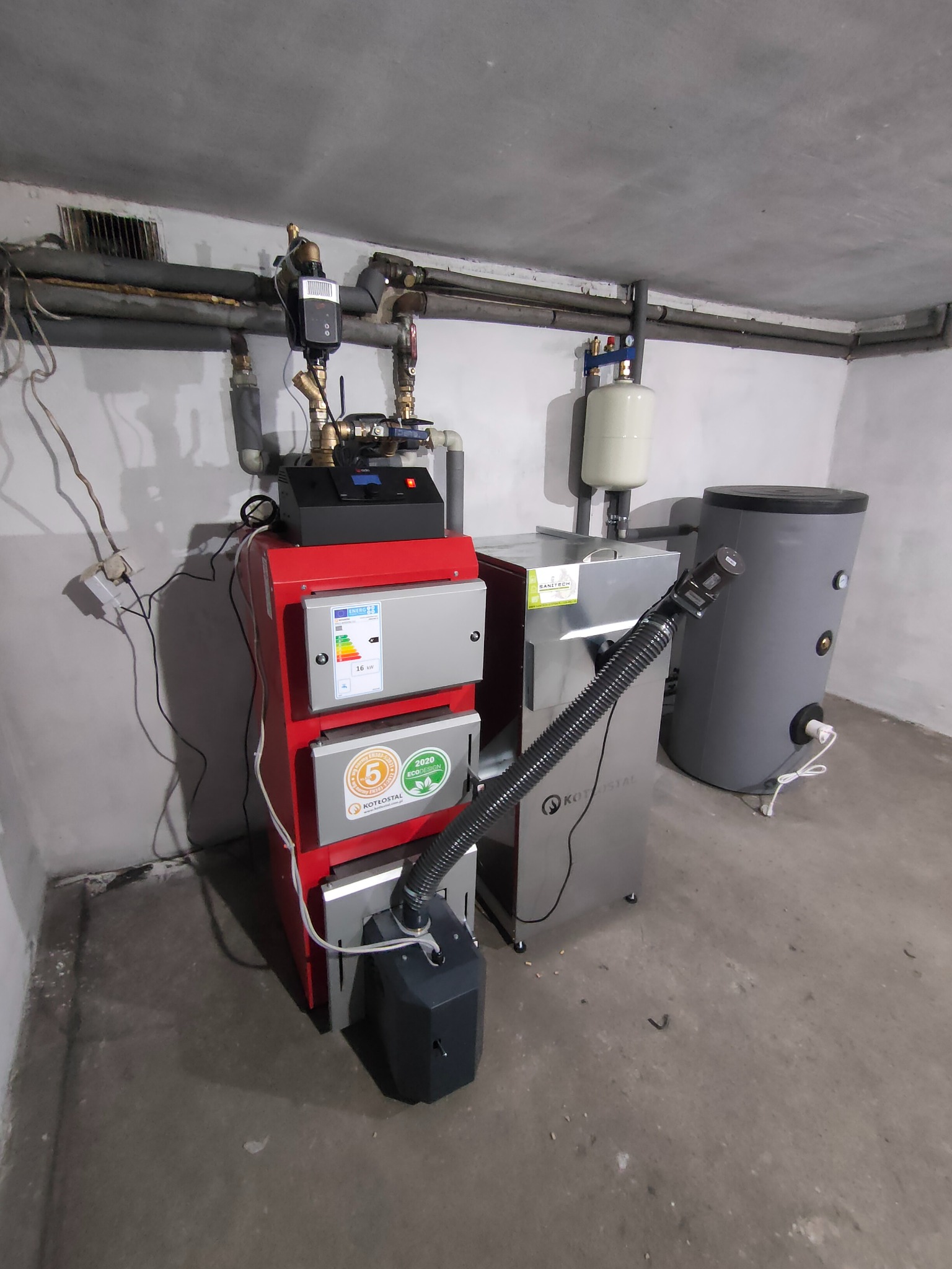 Czerwony piec CO marki Kotlostal z podajnikiem, zbiornikiem na wodę i elementami instalacji grzewczej w szarej kotłowni.