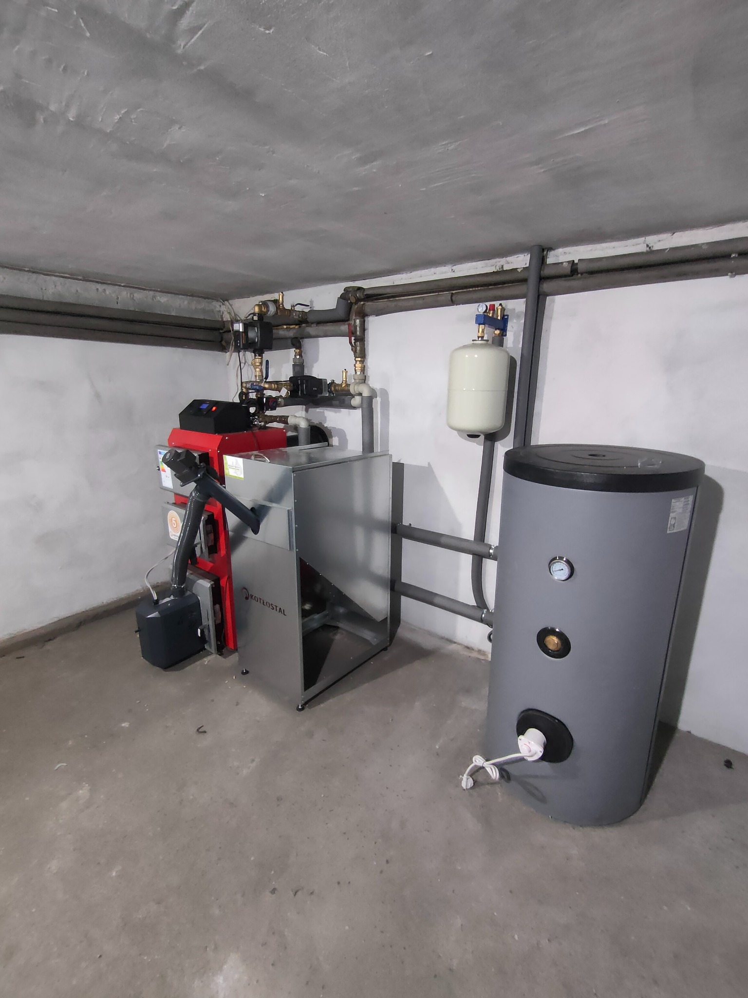 Zainstalowany piec CO marki KOTŁOSTAL z szarym zasobnikiem ciepłej wody w piwnicy z widocznymi rurami grzewczymi pod sufitem.