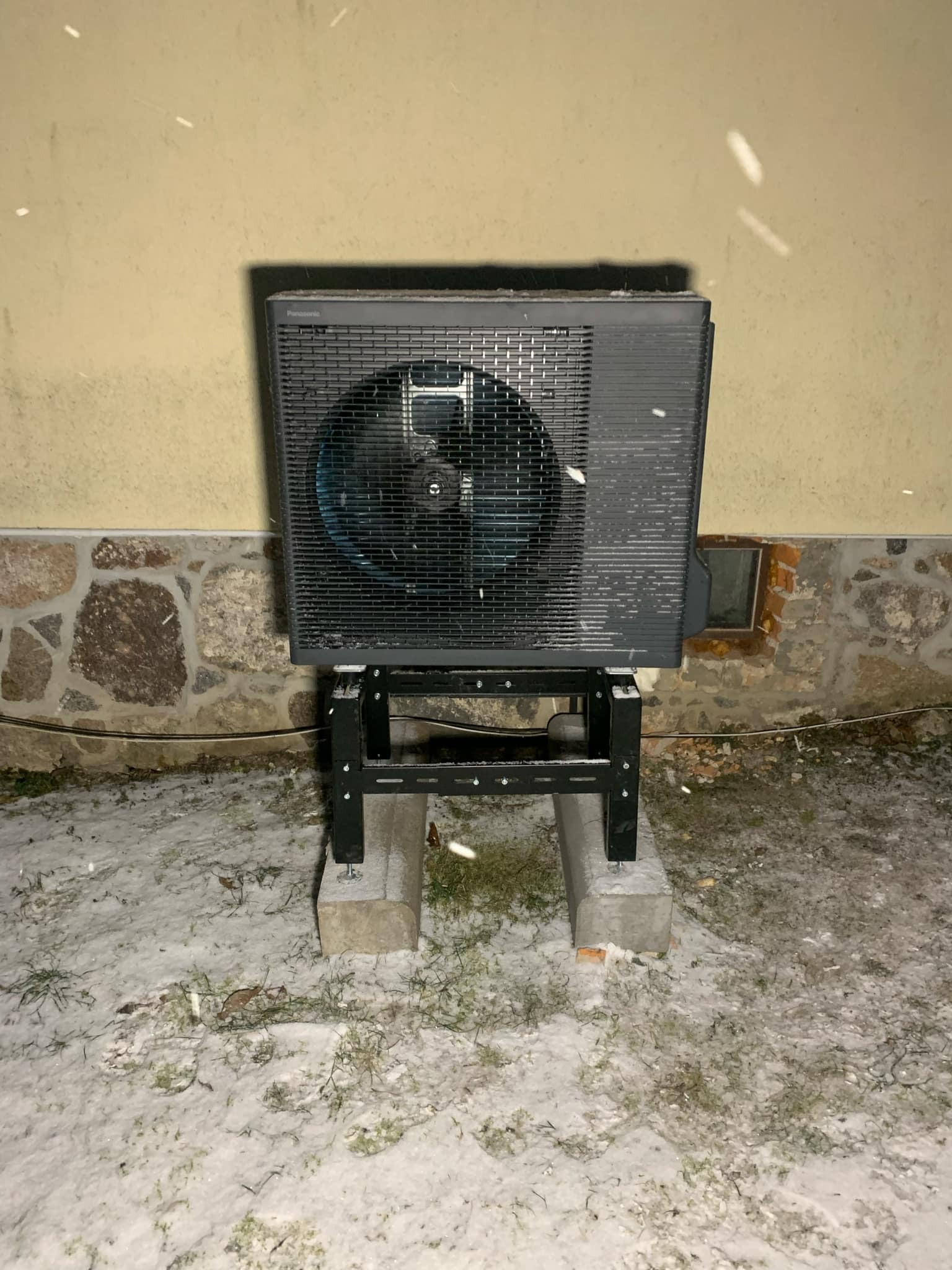 Zewnętrzna jednostka pompy ciepła Panasonic Aquarea zamontowana na metalowej konstrukcji, stojącej na betonowych bloczkach, w zimowej scenerii z padającym śniegiem, na tle ściany budynku...