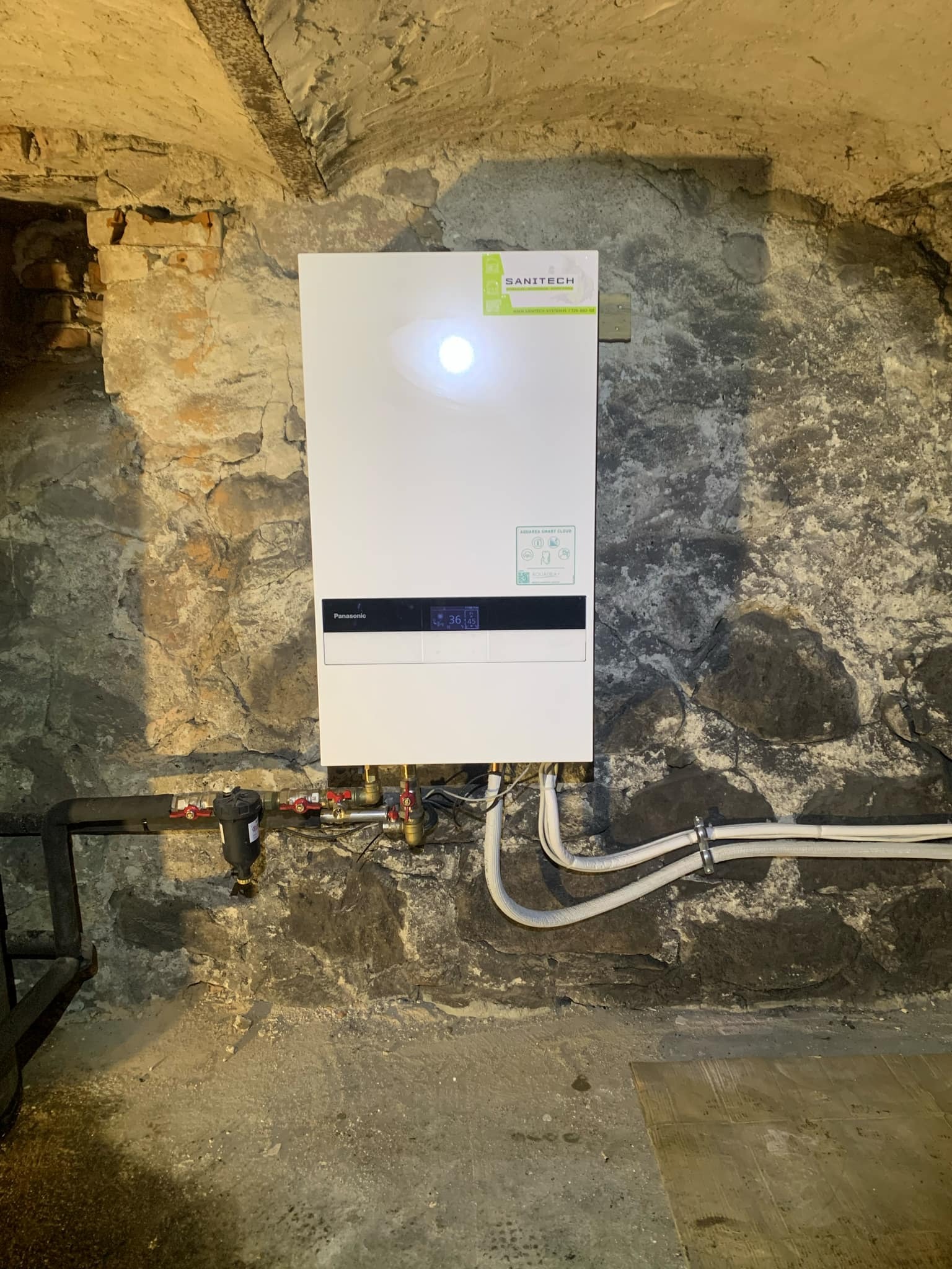 Biała pompa ciepła Panasonic zamontowana na nierównej, kamiennej ścianie piwnicy. Widoczne podłączenia hydrauliczne i elektryczne. Ściana z surowego kamienia z widocznymi spoinami. Niskie...