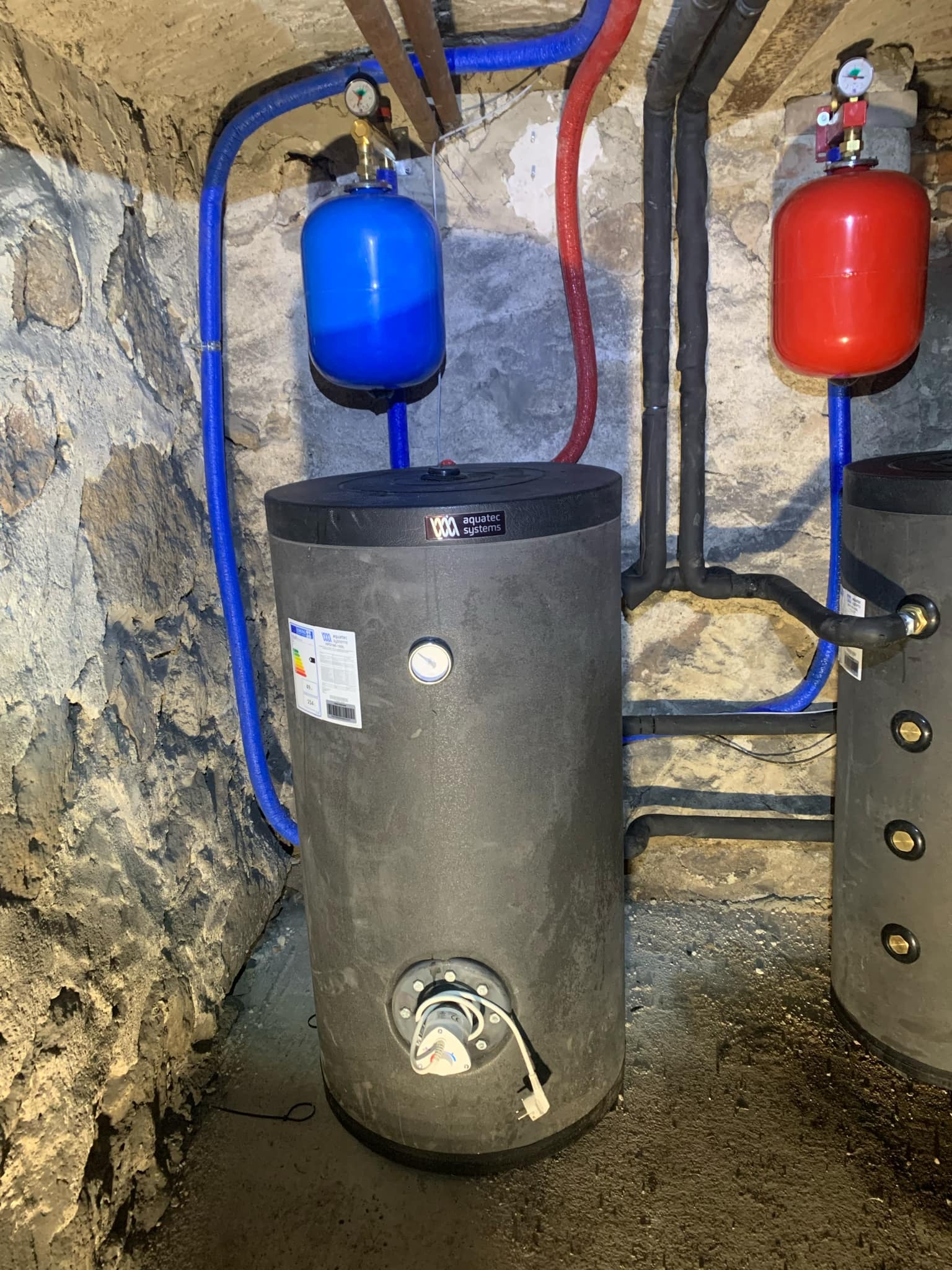 Dwa szare zbiorniki akumulacyjne w piwnicy z kamiennymi ścianami, podłączone do instalacji z niebieskimi i czerwonymi rurami, z widocznymi manometrami i etykietami energetycznymi.