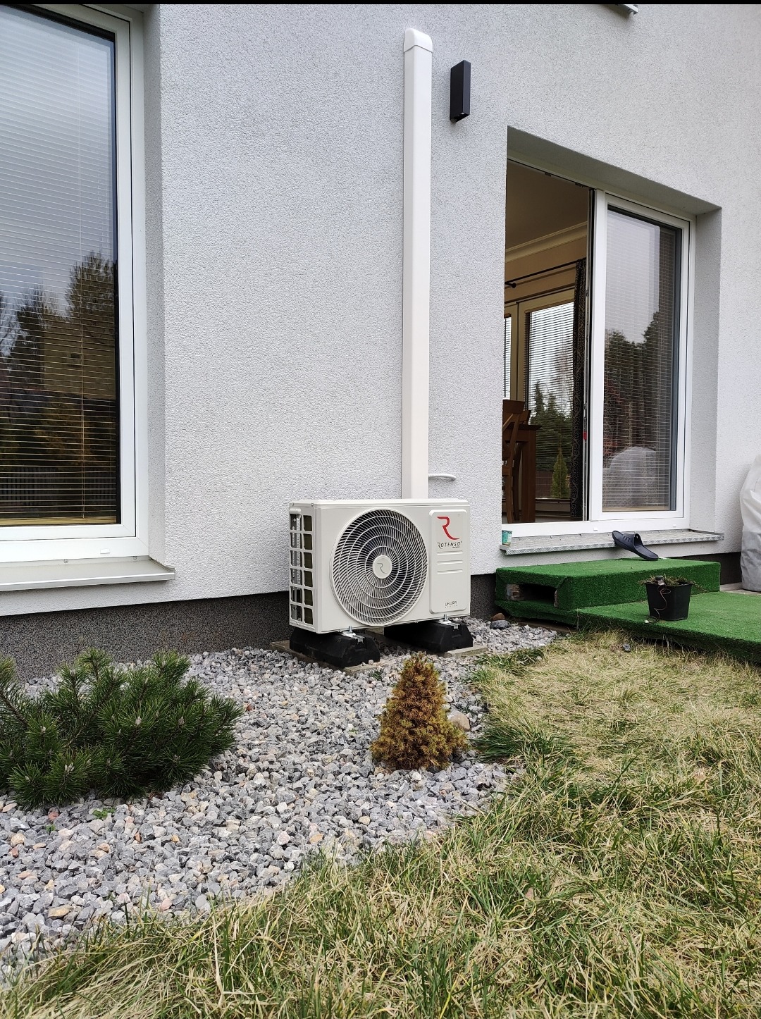 Zewnętrzna jednostka klimatyzacji typu split, marki Rotenso, zamontowana na elewacji domu, z widocznymi przewodami w białej rurze, otoczona ozdobnym kamieniem i małymi krzewami iglastymi. W tle...