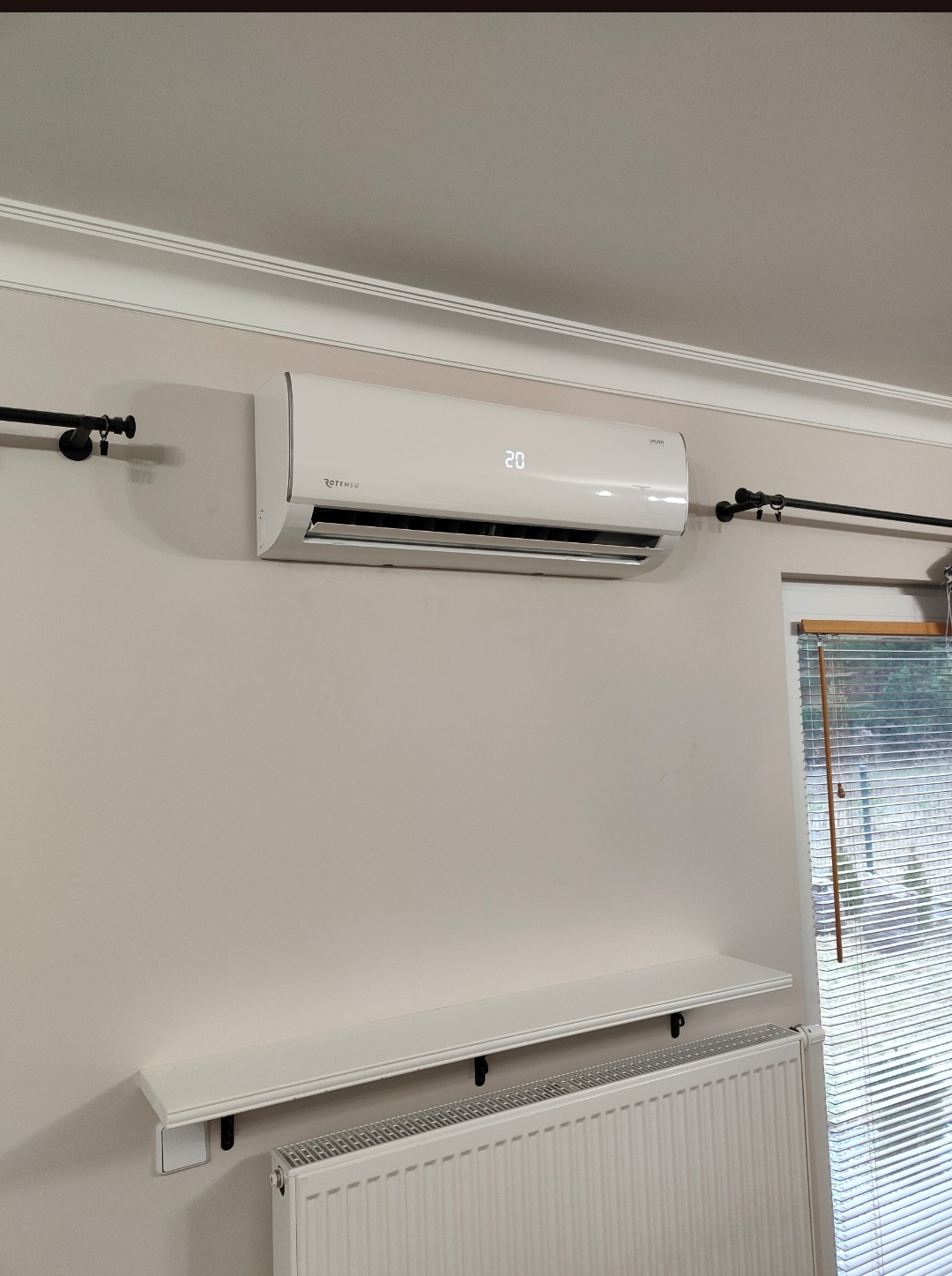 Biały klimatyzator Rotenso zamontowany na ścianie nad grzejnikiem, widoczny wyświetlacz z temperaturą 20 stopni Celsjusza, obok okno z żaluzjami.