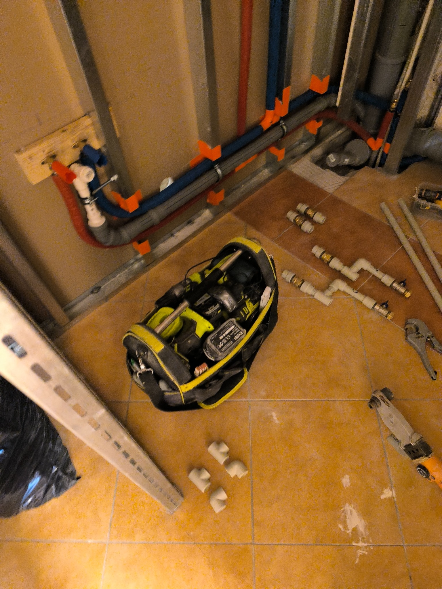 Instalacja hydrauliczna w trakcie montażu: widoczne rury PEX w kolorach niebieskim i czerwonym przymocowane do metalowych profili, torba narzędziowa Ryobi, złączki, rurki i narzędzia na brązowych...