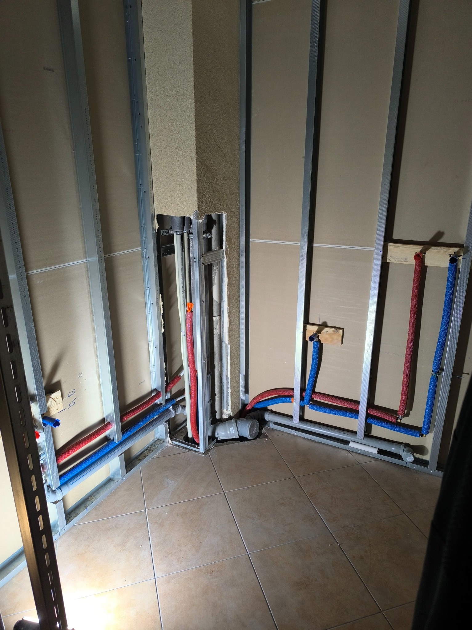 Wnętrze w trakcie prac instalacyjnych: widoczne rury grzewcze (czerwone i niebieskie) prowadzone w metalowym stelażu przy ścianie, przygotowane do zabudowy płytami gipsowo-kartonowymi, z widocznymi...