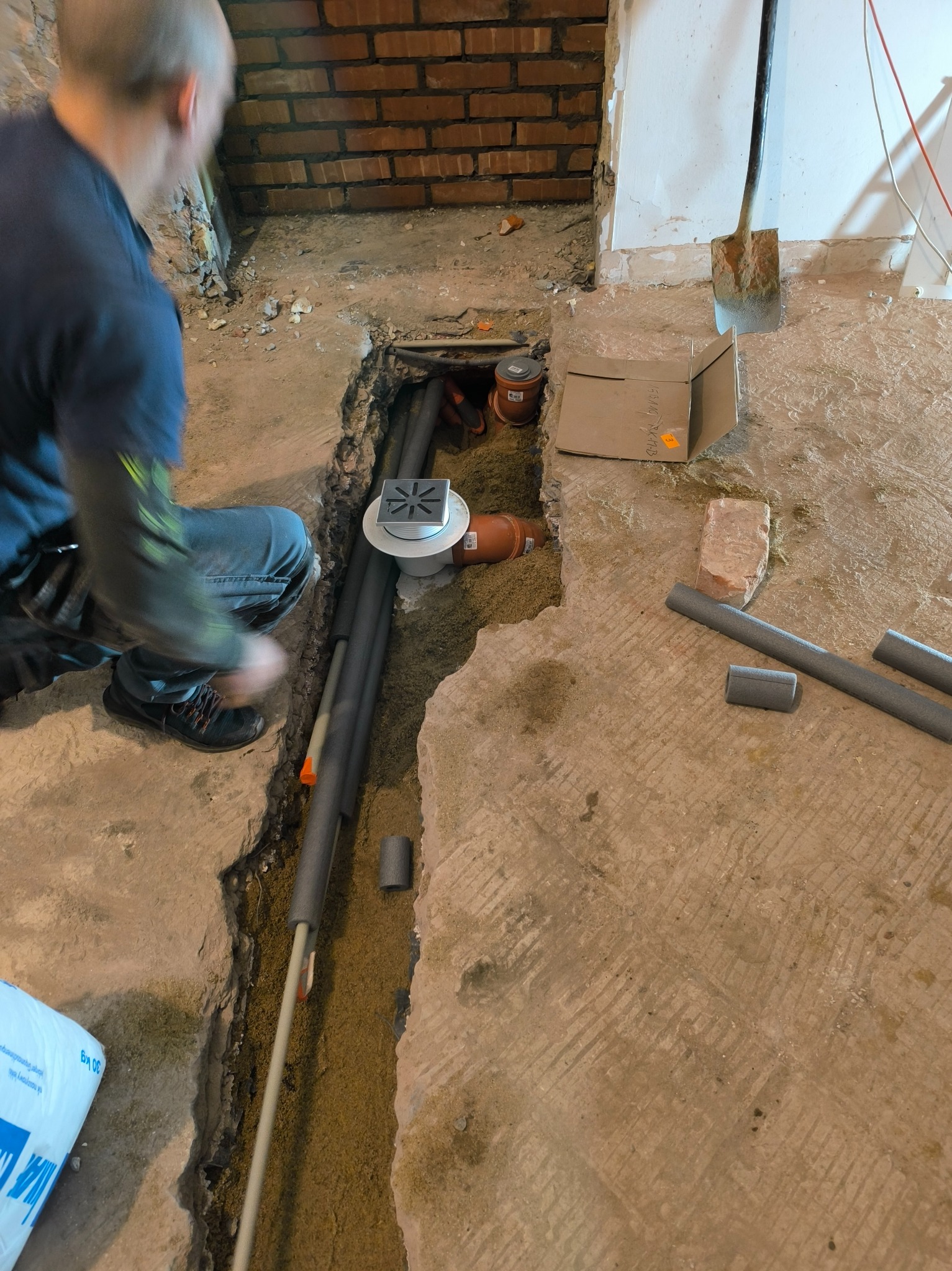 Instalacja odpływu liniowego pod prysznicem: hydraulik w trakcie montażu rur kanalizacyjnych w wykopanym rowie w podłodze, widoczny syfon z kratką i izolowane rury.