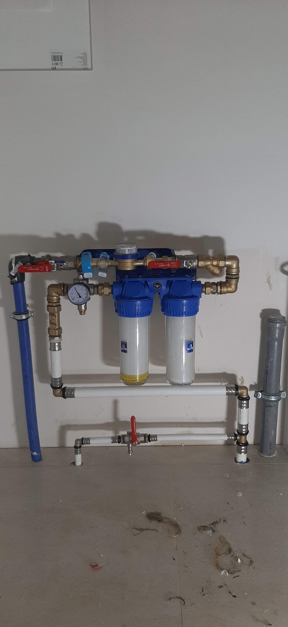 Dwu-stopniowy filtr wody z manometrem, zamontowany na ścianie, z widocznymi rurami doprowadzającymi i odprowadzającymi wodę. W dolnej części widoczny zawór odcinający dopływ wody. Na podłodze...