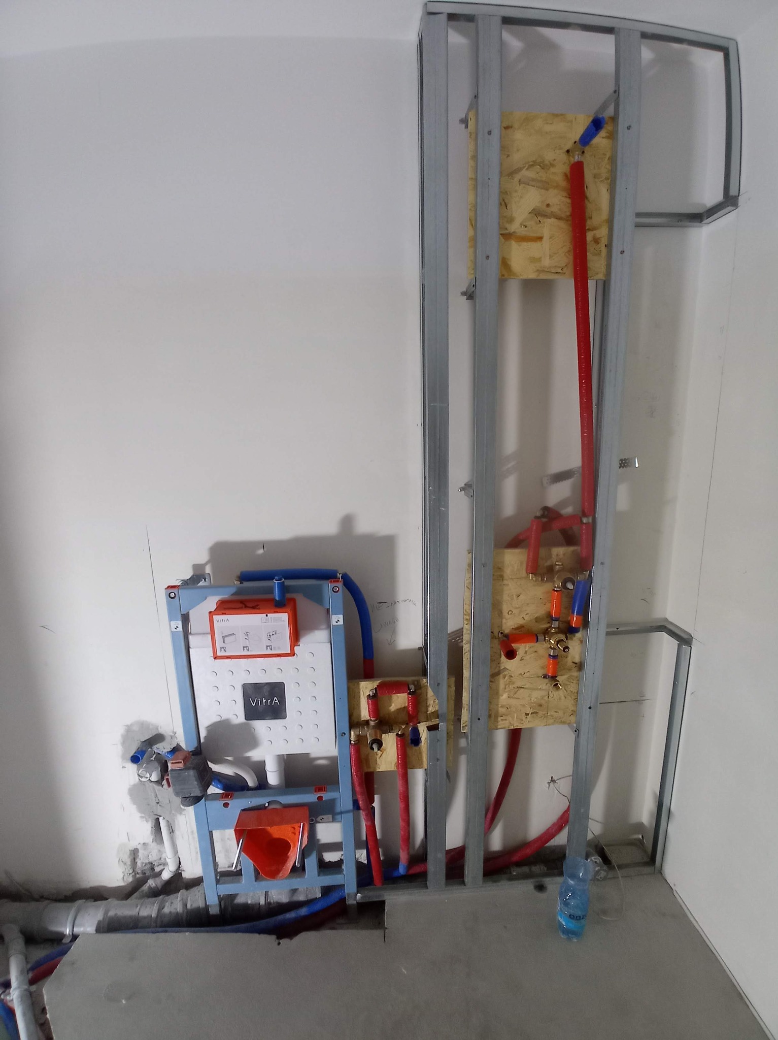 Stelaż podtynkowy WC marki Vitra zamontowany w konstrukcji z profili, widoczne podłączenia hydrauliczne z rur PEX w kolorach czerwonym i niebieskim, oraz butelka wody na podłodze.