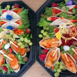 Dwie czarne tace z cateringiem: tortille z warzywami i mięsem, kawałki kurczaka w sosie, winogrona i truskawki. Dekoracje 'Happy Birthday'.