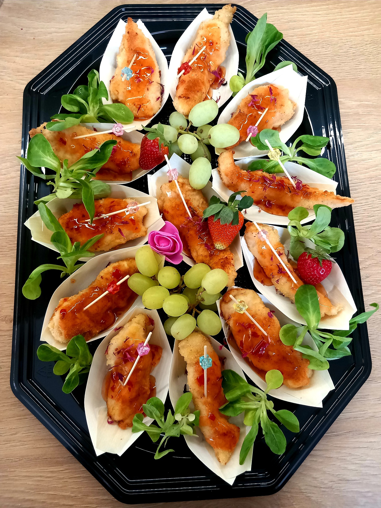 Czarny, ośmiokątny talerz z przekąskami: smażone kawałki mięsa w łódeczkach z papieru, polane sosem, udekorowane sałatą, winogronami i truskawkami na wykałaczkach.